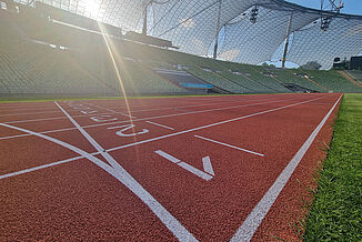 Tiefwinkelaufnahme der roten Kunststofflaufbahn im Olympiastadion München bei Sonnenschein. Im Vordergrund ist deutlich die Ziellinie mit Bahnenmarkierung zu erkennen – Symbol für Zielankünfte bei Leichtathletikrennen.
