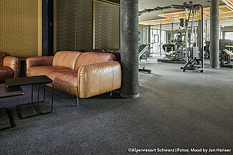Trainingsbereich und abgetrennte Sitzecke. Sitzbereich mit braunen Sofas in einem Fitnessstudio, im Hintergrund sind Trainingsgeräte zu sehen. Der Bodenbelag ist dunkelgrau.