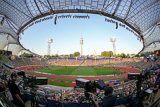Innenansicht des Olympiastadions München während der Leichtathletik-Europameisterschaft 2022. Die rote Kunststofflaufbahn und das zentrale Spielfeld sind vorbereitet für Wettkämpfe. Kameras und Publikum zeigen die Live-Atmosphäre dieses internationalen Events.