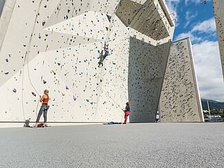 Outdoor-Kletterbereich mit Fallschutzboden. Der Bodenbelag ist grau. An der Kletterwand sind einige kletternde Menschen zu sehen, auf dem Boden stehen die Sicherungspartner. 