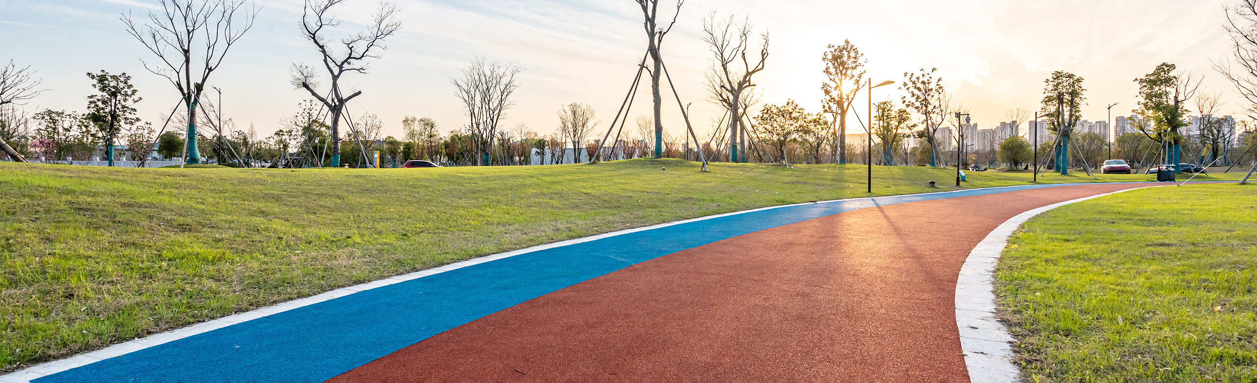 Synthetischer Jogging-Track im Freien, in Rot-Blau-Farbgebung, eingebettet in eine grüne Parklandschaft bei Sonnenuntergang. Der Boden besteht aus elastischem, wasserdurchlässigem Kunststoffbelag – ideal für gelenkschonendes Lauftraining.