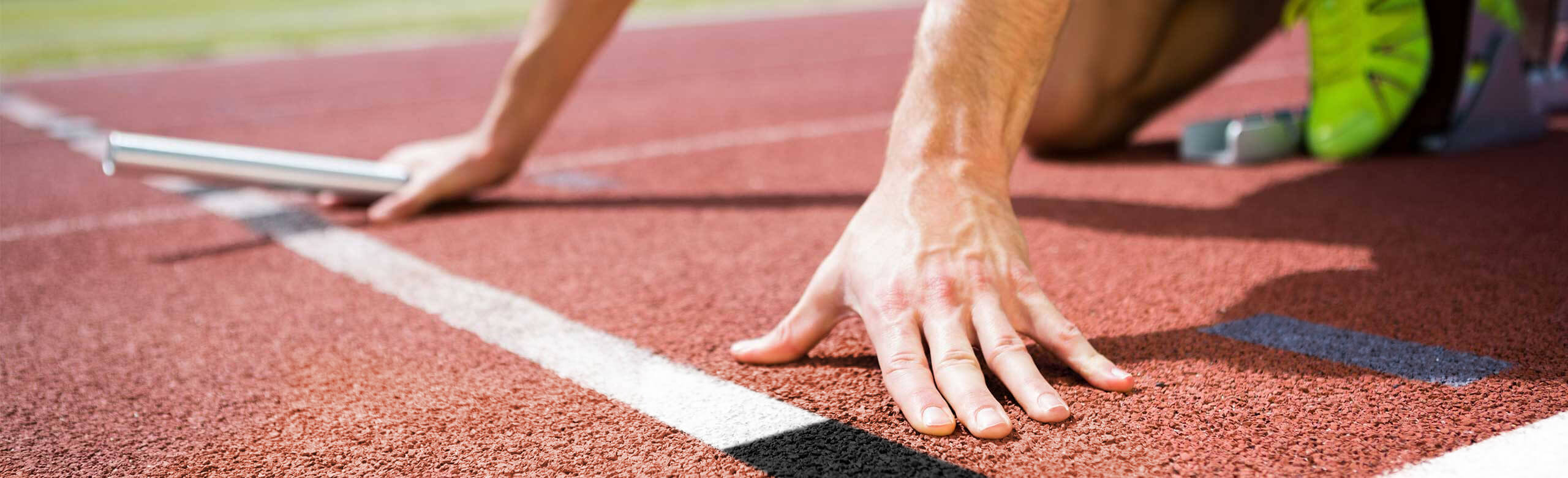 Close-up of an athlete in starting position on a red synthetic track, holding a relay baton, with starting blocks and spikes.