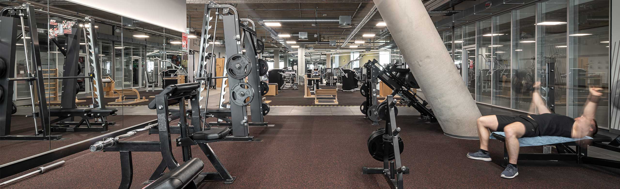 REGUPOL Bodenbelag in einem Bereich für Freihanteltraining in einem Fitnessstudio. REGUPOL Bodenbelag in einem Bereich für Freihanteltraining in einem Fitnessstudio. Man sieht einen Mann liegend auf einer Hantelbank beim Gewichtheben.