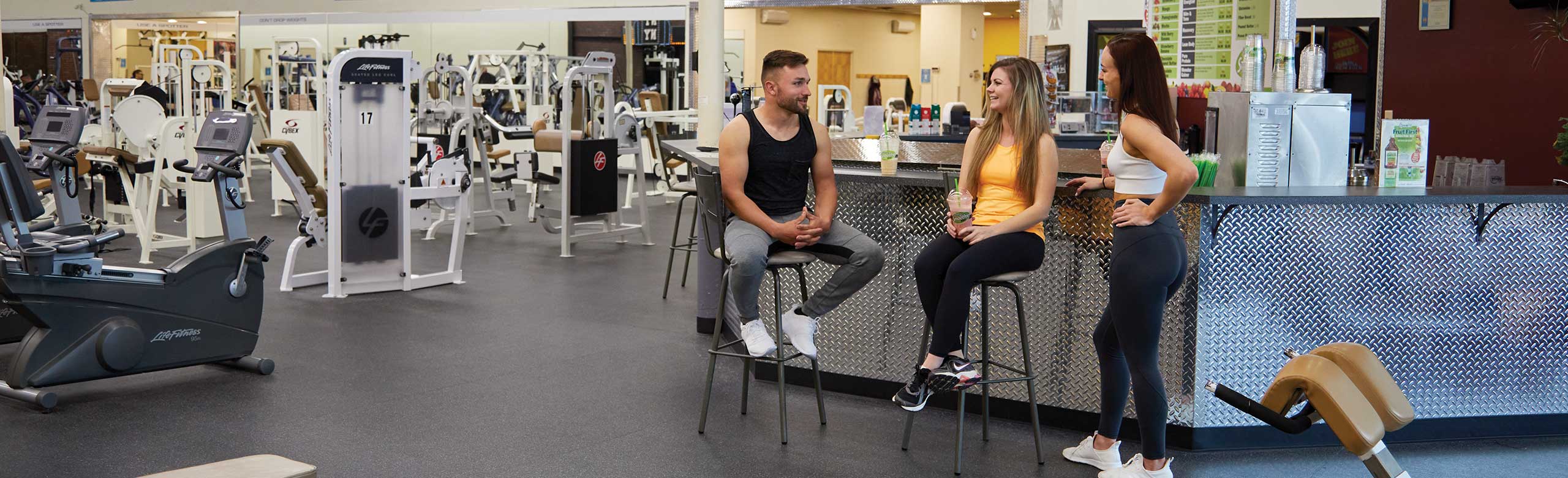 Zwei Frauen und ein Mann unterhalten sich an einer Theke im Fitnessstudio. Zwei Frauen und ein Mann unterhalten sich an einer Theke im Fitnessstudio. Im Hintergrund sind Fitnessgeräte zu sehen. Der REGUPOL Bodenbelag ist im dunklen Grau gewählt worden.