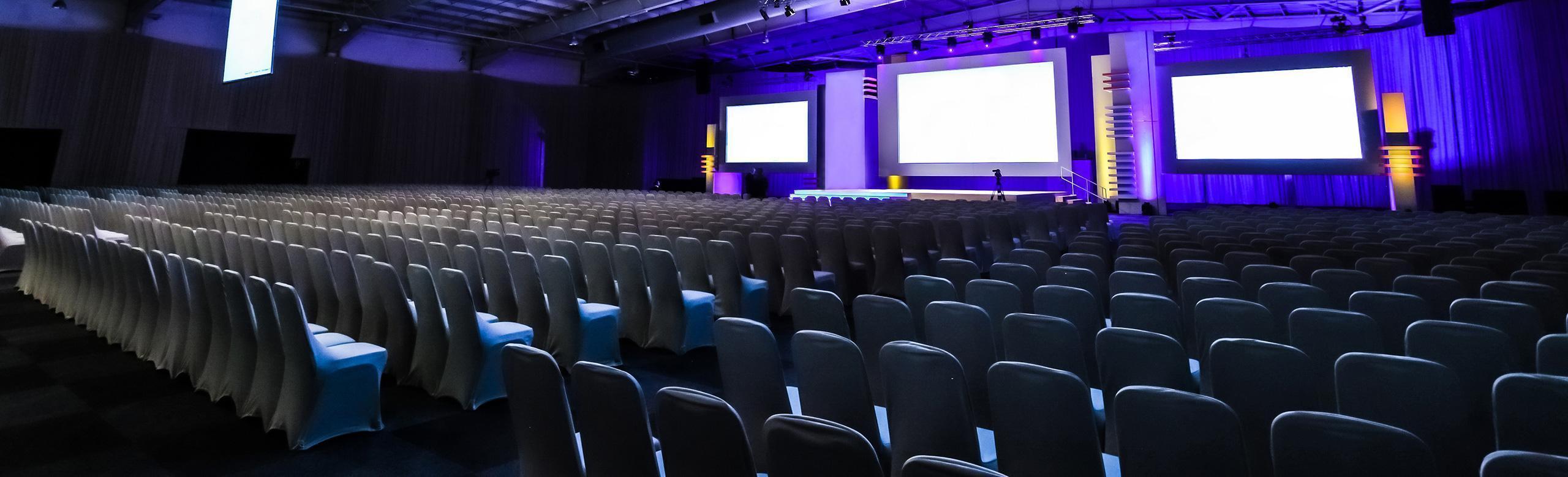 Viele Stuhlreihen, die in Richtung Bühne weisen.  Die Bühne befindet sich in einer Sporthalle und ist mit lilafarbenem Licht hinterleuchtet. Drei große Bildschirme hängen an der Wand der Bühne. 