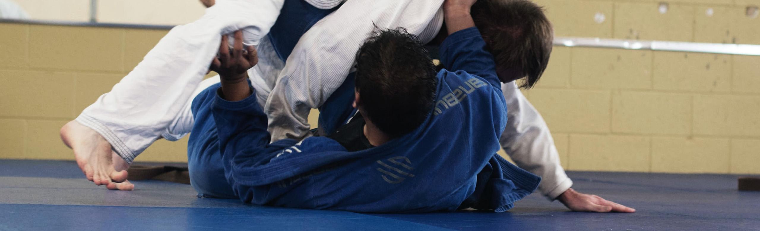 Zwei Personen tragen Jiu-Jitsu-Anzüge und befinden sich im Bodenkampf auf blauen Matten in einer Sporthalle