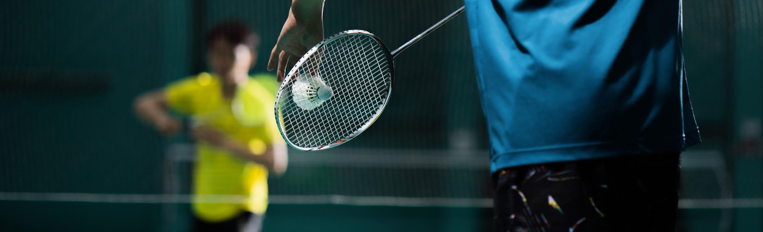 Zwei Personen spielen Badminton in einer Sporthalle. Im Vordergrund ist ein Spieler mit Schläger und Federball zu sehen, im Hintergrund steht die zweite Person spielbereit.