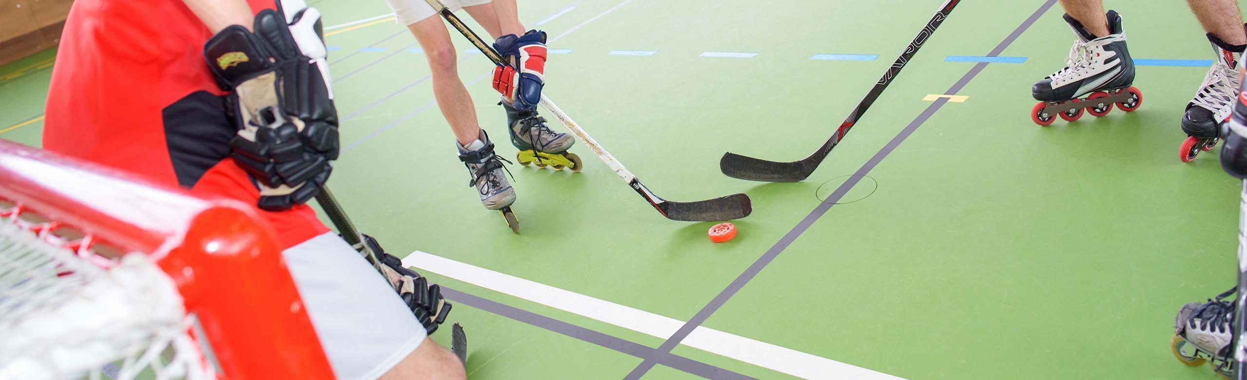 Rollsport in einer Sporthalle Verschiedene Personen spielen Inline-Hockey in einer Sporthalle auf einem grünen Sporthallenboden mit verschiedenen Linierungen. Sie haben Inline-Skates und Hockeyhandschuhe an, Hockeyschläger in den Händen und spielen sich den Puck vor einem Tor zu.