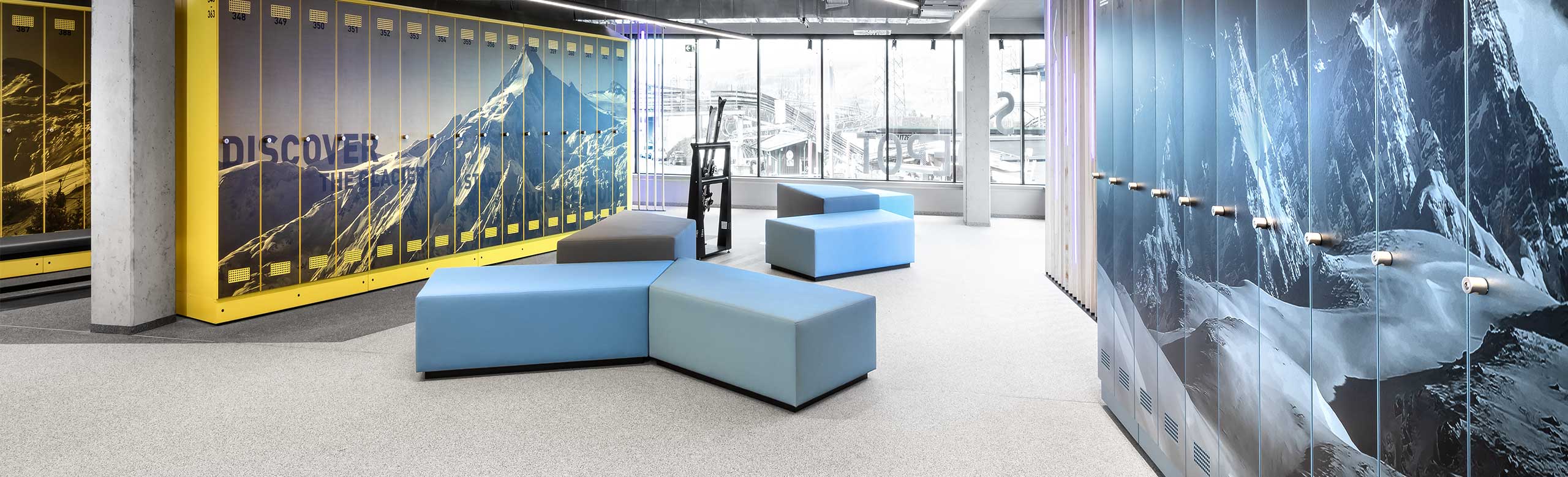 Ein helles Skidepot in einer Bergbahnstation. Am Ende des Raumes ist eine große Fensterfront, rechts und links des Bildes sieht man Spinde mit Berg-Motiven. In der Mitte befinden sich mehrere Sitzgelegenheiten in Blau und Grau. Der Bodenbelag ist hell, unterhalb der Spinde ist er dunkelgrau. 